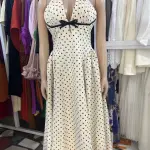 Vintage-inspired polka dot halter-neck maxi dress with black polka dots, a plunging neckline, and a contrasting black bow or ribbon accent at the bust.